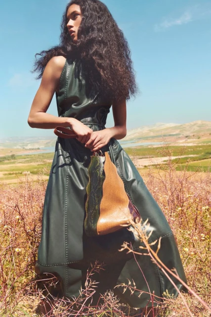 Fashion shot of woman in a green dress holding a leather bag in a sunlit, golden field against a clear blue sky. Low angle, eye-level view.