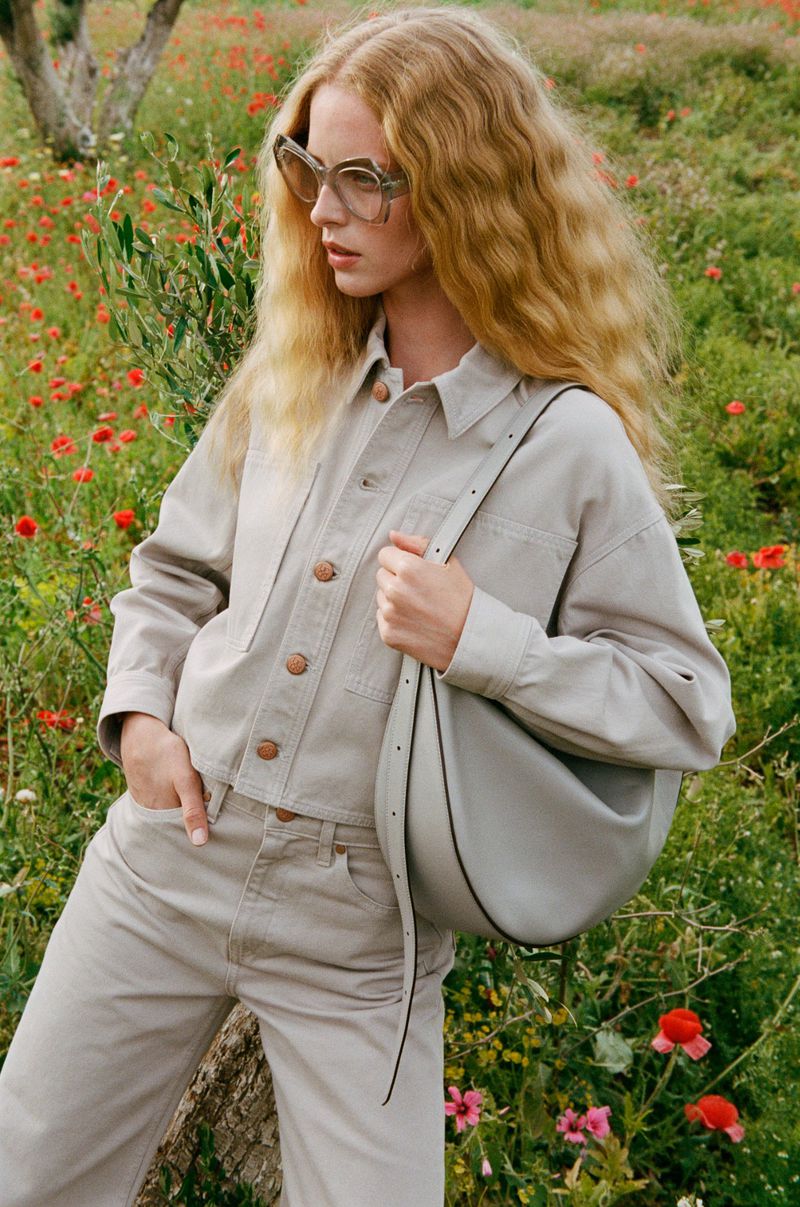 Blonde woman in beige denim jacket and pants stands in a field of red poppies, carrying a gray bag, and wearing sunglasses.