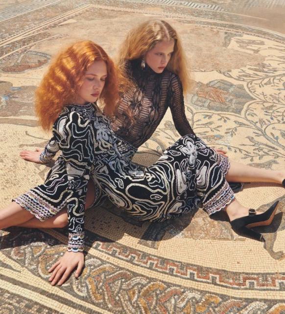 Two red-haired women pose on a mosaic-tiled surface, wearing patterned dresses, against a sunlit, textured backdrop in an outdoor setting.