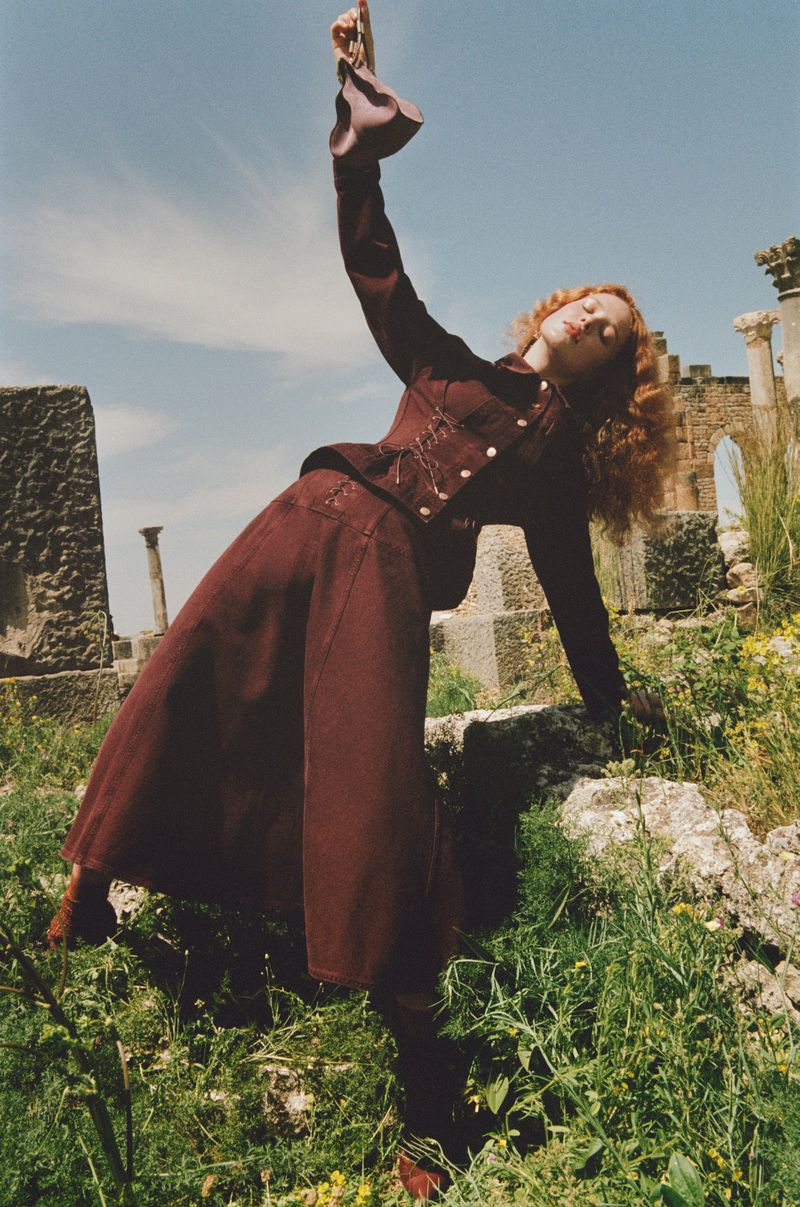 Fashion shot of a woman in a maroon skirt suit leaning back on a stone wall surrounded by green vegetation under a bright sky.