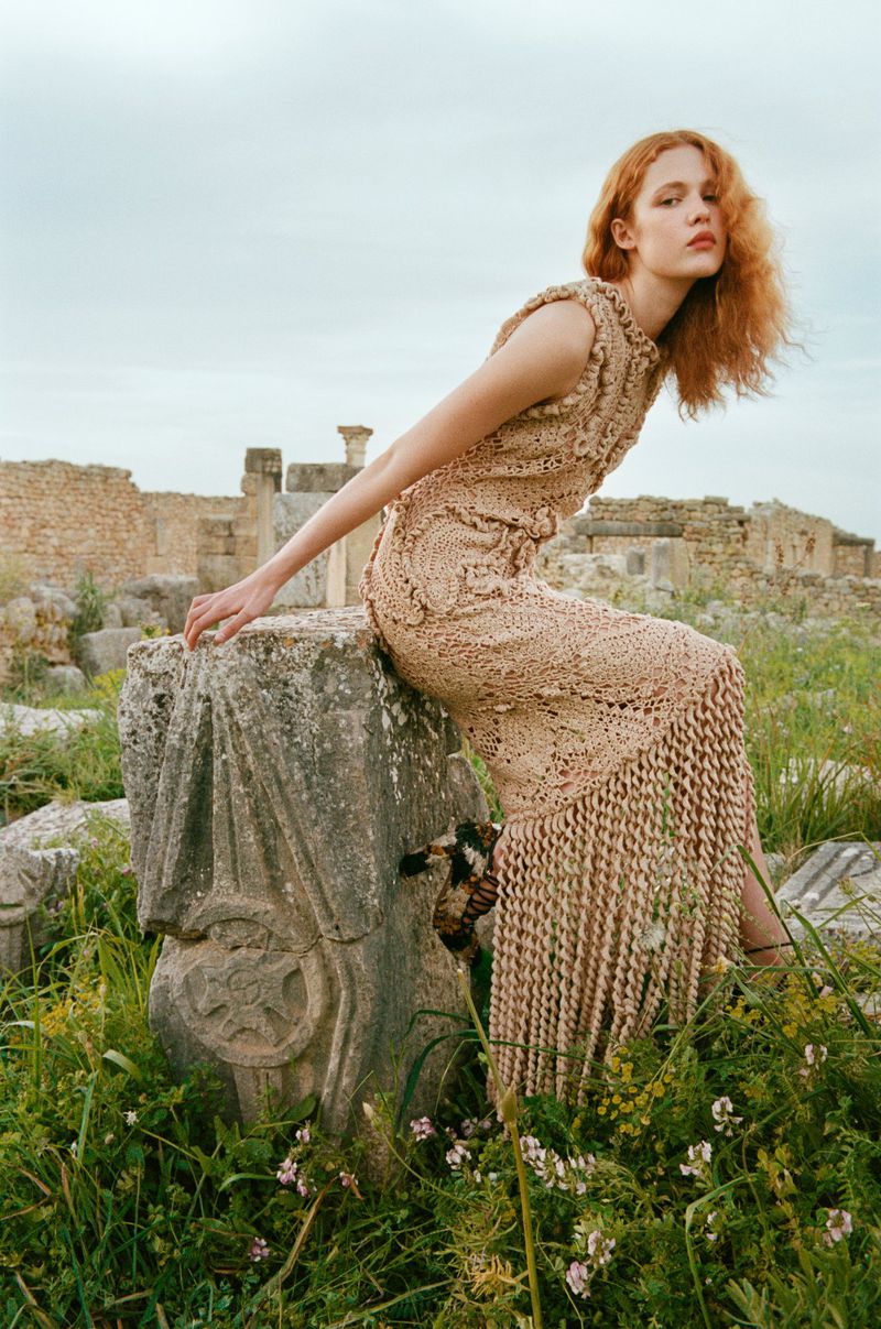 A red-haired woman in an ornate dress sits on a stone ruin. Overcast light illuminates a grassy field and the weathered stones behind her.