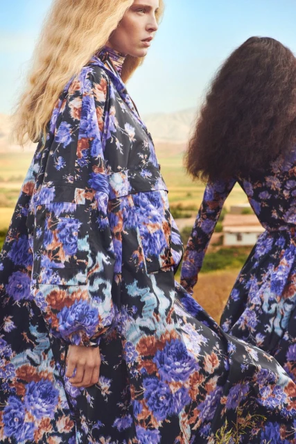 Two women wearing floral print dresses in a field. One woman has blonde curly hair and the other has dark curly hair.