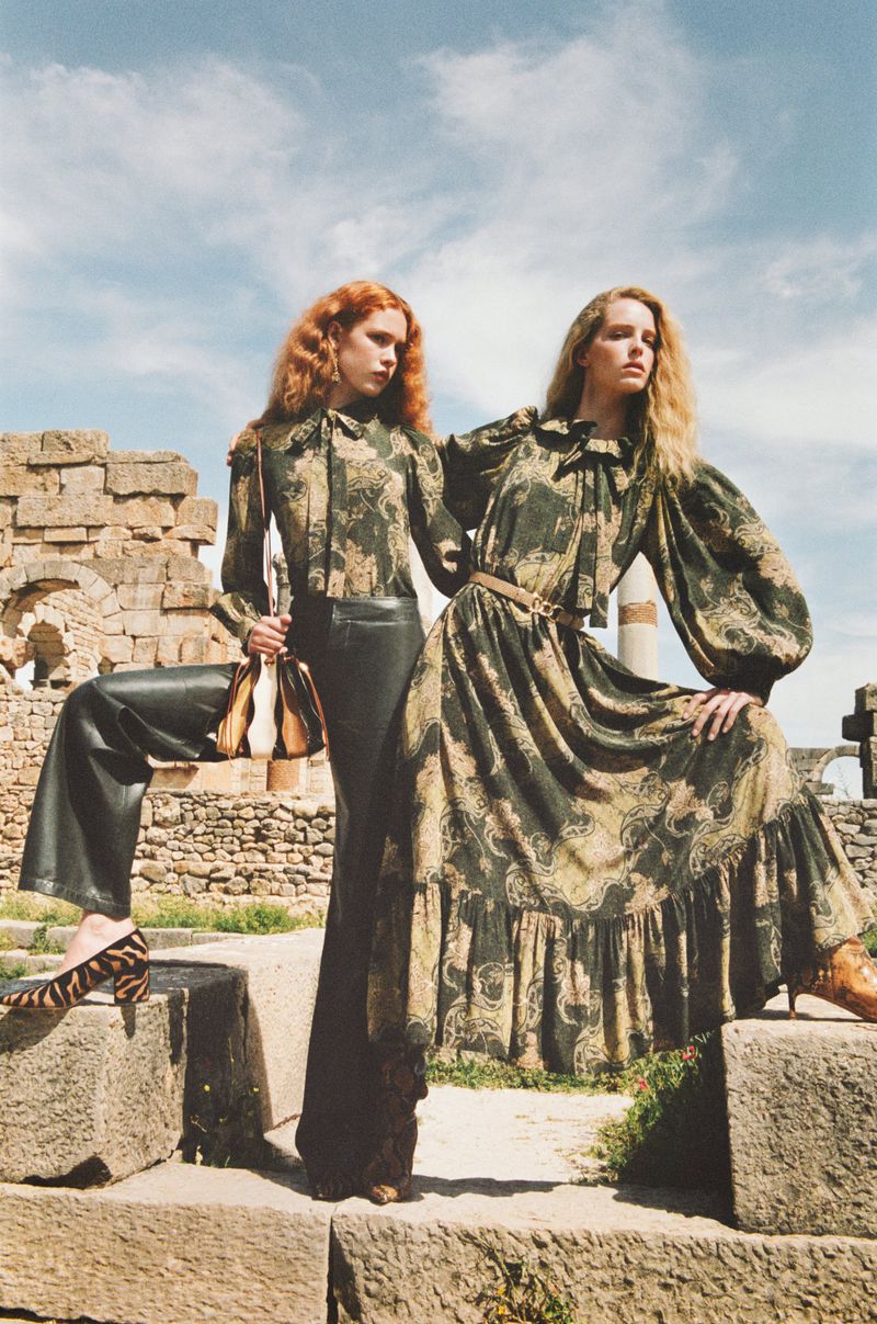 Two women in patterned tops and dark skirts pose in the ruins, one with a purse and animal print shoes, under a bright sky.