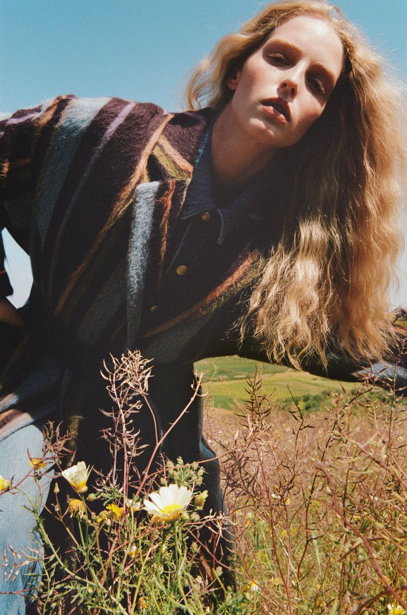 Fashion shot of a blonde woman wearing a striped coat in a rural field of flowers under a blue sky in a medium outdoor shot.