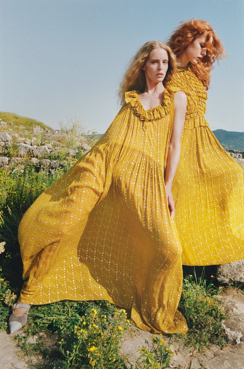 Two women in mustard yellow dresses posing outdoors on a sunny day in a grassy, rocky field with a clear blue sky above them.