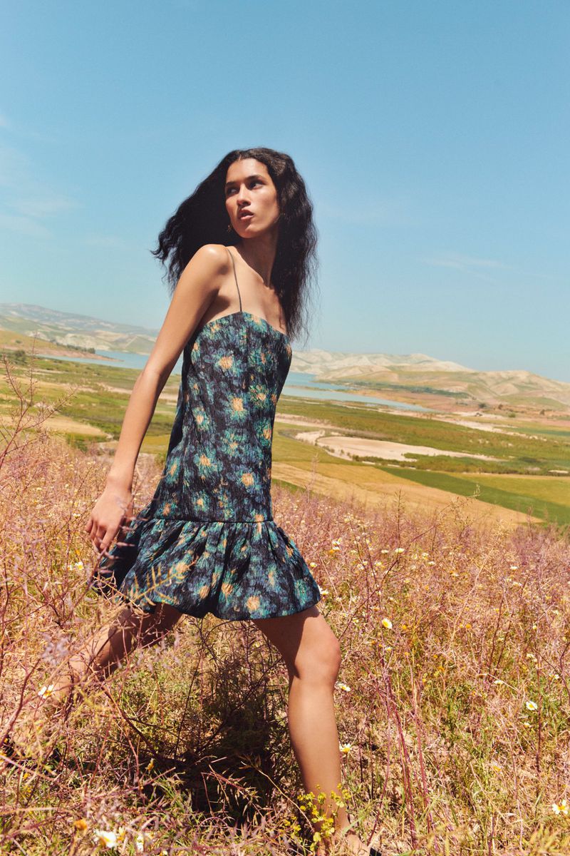 A woman in a patterned dress walks through a sunlit field with a bright blue sky and distant hills.