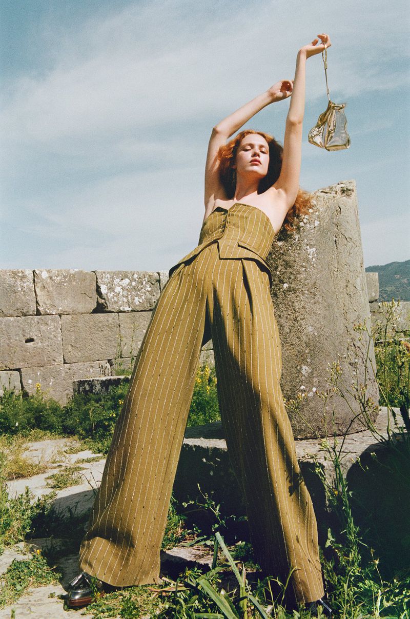 Fashion shot: A red-haired woman in a two-piece olive, pin-striped suit poses near ancient ruins, holding an ornate, gilded lantern.