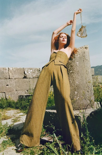 Fashion shot: A red-haired woman in a two-piece olive, pin-striped suit poses near ancient ruins, holding an ornate, gilded lantern.