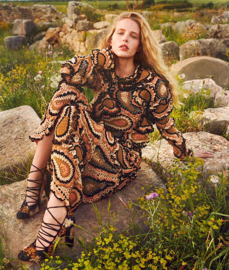 Fashionable blonde model poses in a crocheted paisley dress and heels amidst a sunny field with stone ruins, creating a timeless editorial shot.