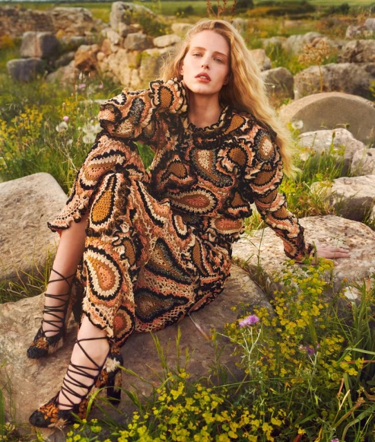 Fashionable blonde model poses in a crocheted paisley dress and heels amidst a sunny field with stone ruins, creating a timeless editorial shot.