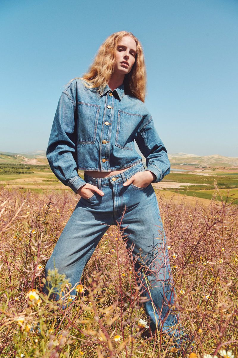 Blonde fashion model poses in blue denim outfit outdoors in a field under a clear sky on a sunny day with a wide shot.