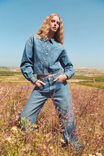 Blonde fashion model poses in blue denim outfit outdoors in a field under a clear sky on a sunny day with a wide shot.