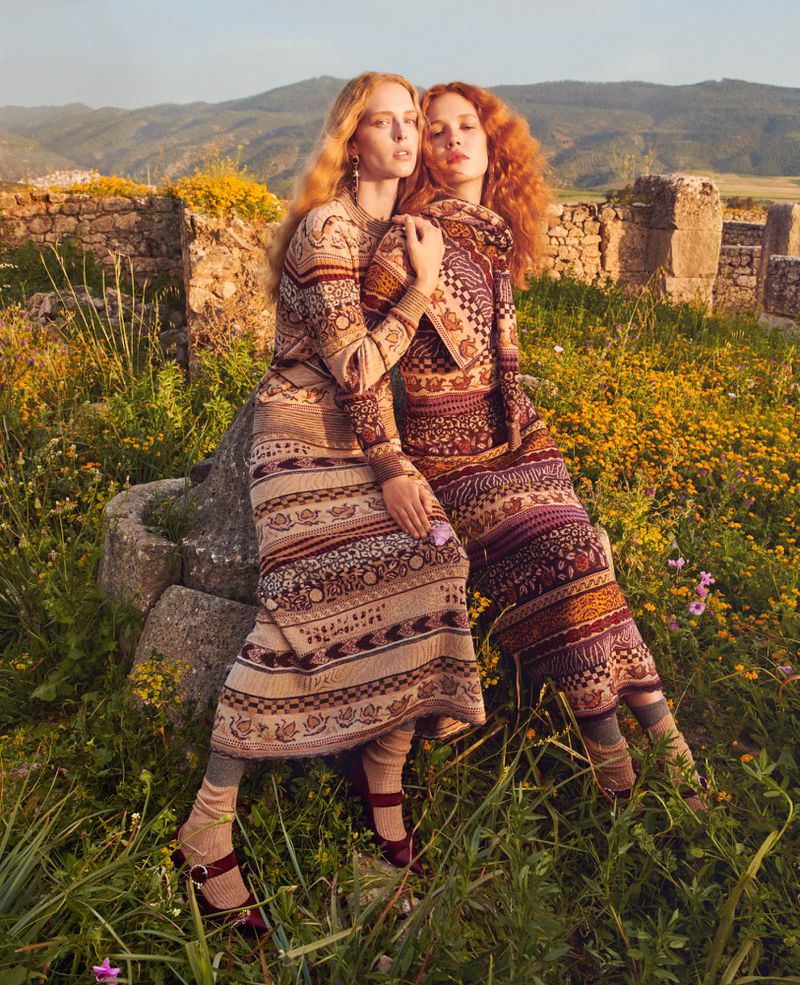 Two women in patterned dresses pose amongst wildflowers and ancient ruins in a fashion editorial shot during golden hour light.