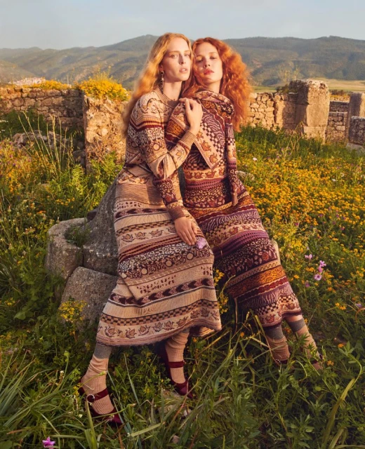 Two women in patterned dresses pose amongst wildflowers and ancient ruins in a fashion editorial shot during golden hour light.