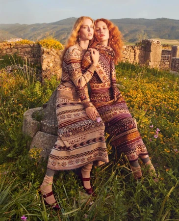 Two women in patterned dresses pose amongst wildflowers and ancient ruins in a fashion editorial shot during golden hour light.