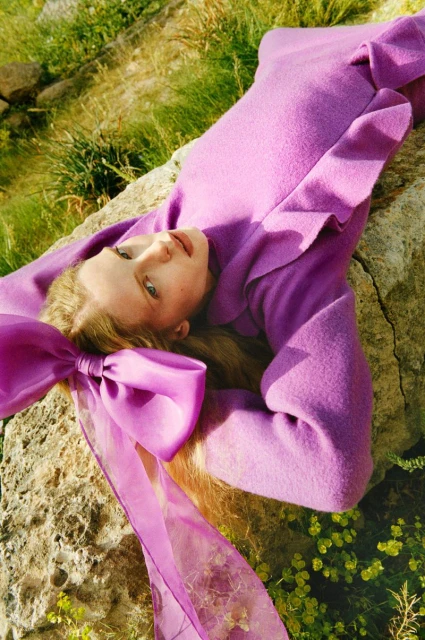 Woman in purple dress with bow lies on rock in sunny field, fashion editorial style. Soft natural light illuminates the outdoor setting.