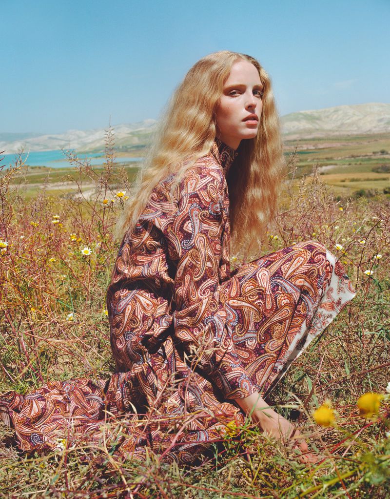 A woman with long blonde hair in a paisley dress is crouched in a grassy field on a sunny day with a lake and hills in the background.