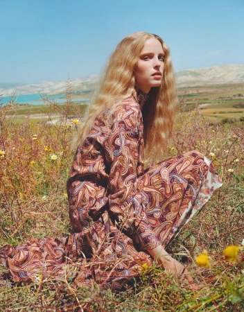 A woman with long blonde hair in a paisley dress is crouched in a grassy field on a sunny day with a lake and hills in the background.