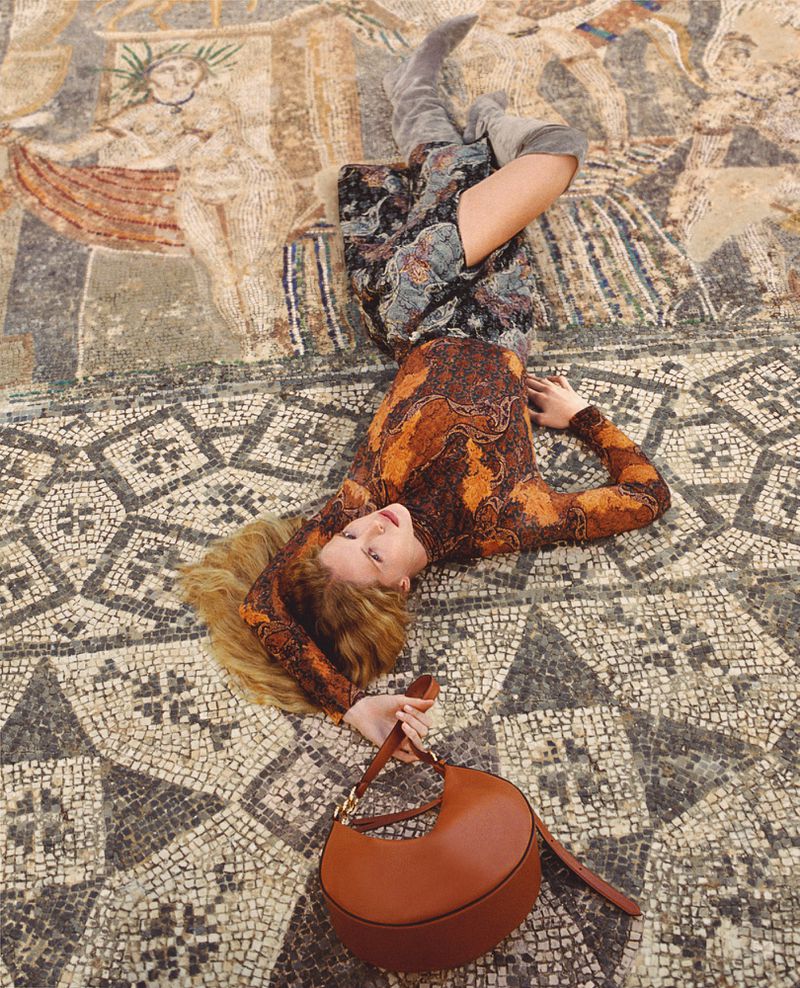 Woman in patterned dress and gray boots poses on mosaic tile floor, holding a brown leather purse in a fashion editorial shot.