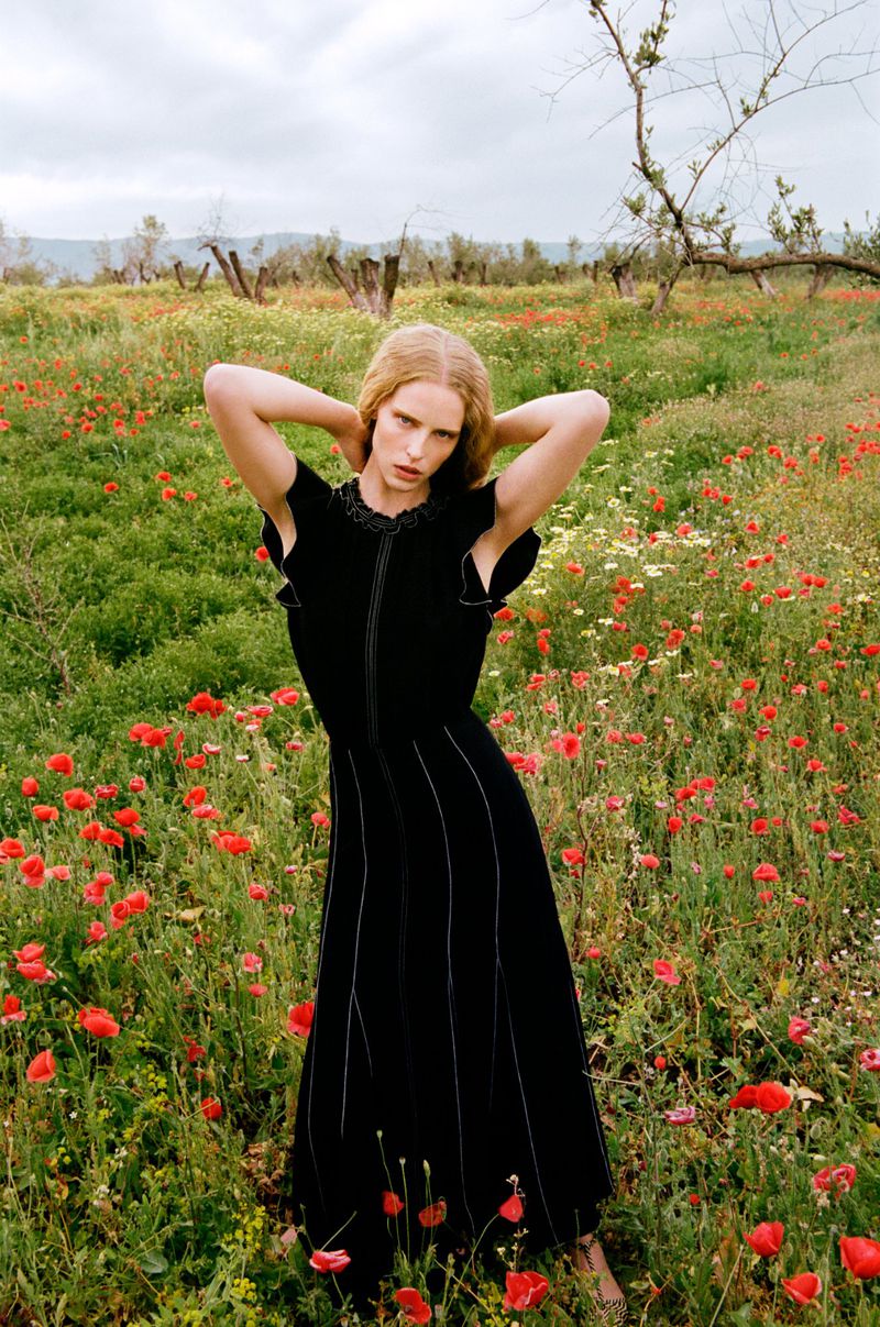 Woman in black dress stands in a field of red poppies and white flowers under an overcast sky. Fashion shot in a rural landscape.