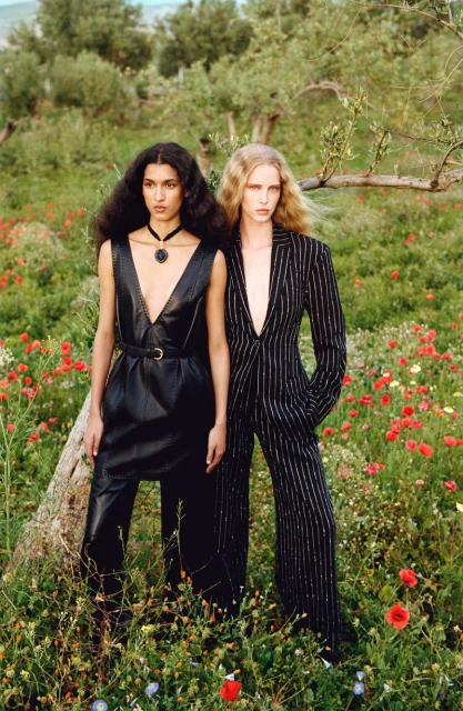 Two women in fashionable attire pose in a field of wildflowers against a backdrop of olive trees and a soft, diffused natural light.