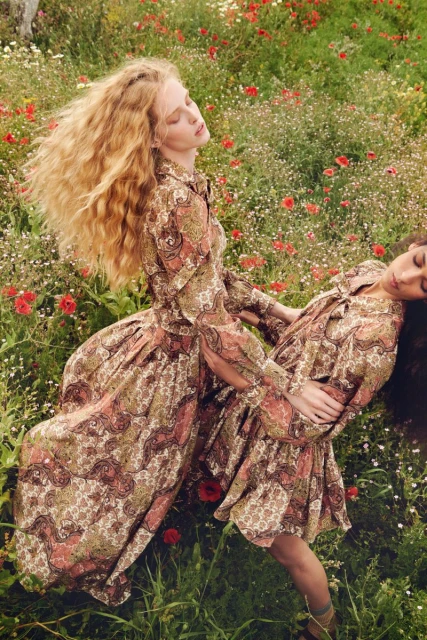 Two women in matching patterned dresses pose in a field of wildflowers and tall green grass, bathed in soft, diffused natural light.