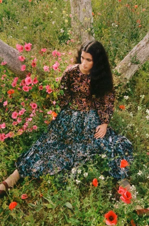 Adult female in a brown and floral long sleeve top and long patterned skirt sitting in a field of wildflowers and tall grass.