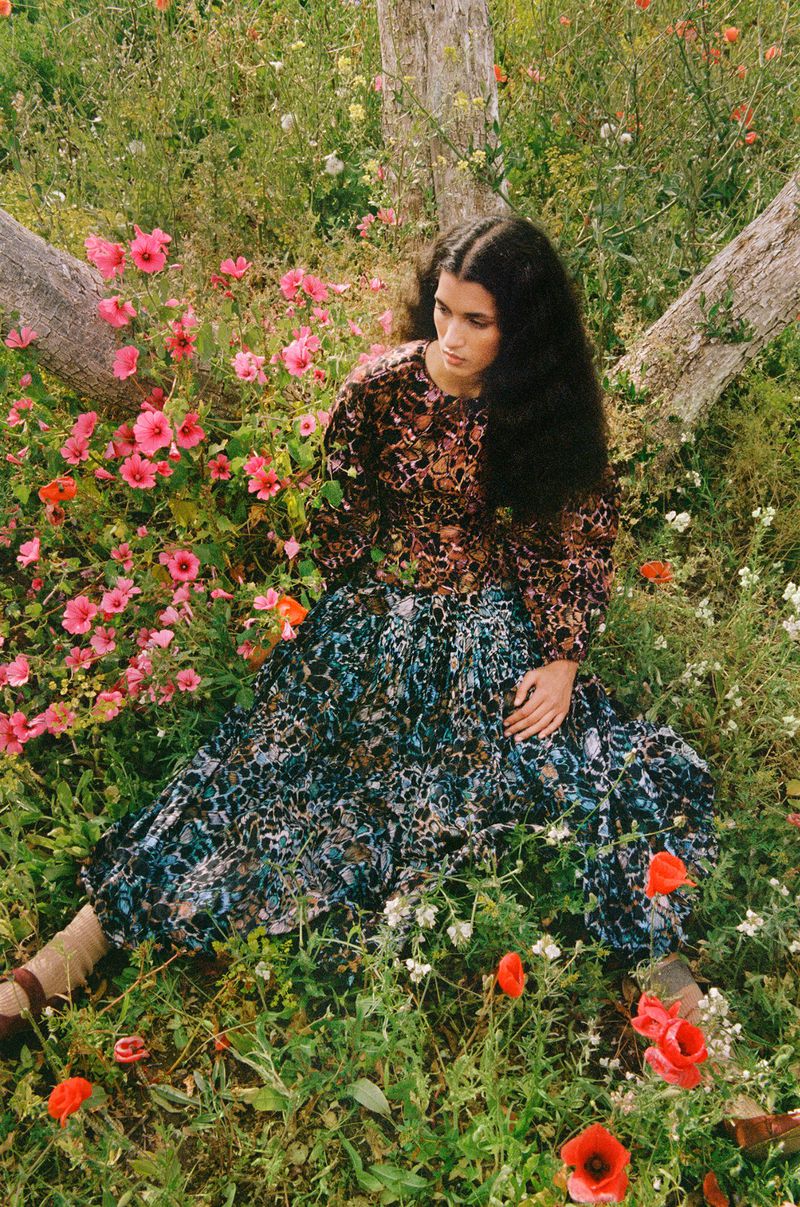 A woman with long dark hair is sitting in a field of flowers wearing a patterned blouse and skirt. Surrounded by lush greenery and blossoms.