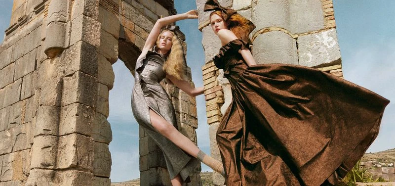 Two women in brown and silver gowns with large hair bows pose beside stone ruins under a blue sky in a fashion editorial.