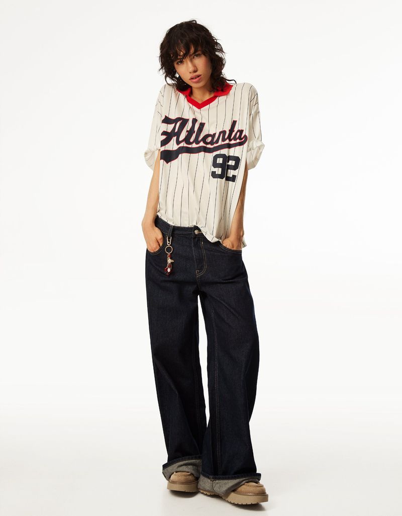 Young woman in white baseball jersey with 'Atlanta 92' paired with dark denim jeans, posing against a clean white backdrop.
