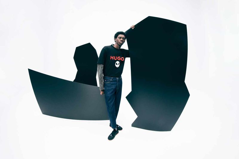 Young man poses in a black t-shirt with a Hugo logo, blue jeans, and black shoes, standing on a white background surrounded by geometric shapes.