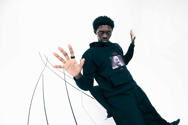 Man in black hoodie and sweatpants strikes a pose in a bright studio, hands raised towards camera, minimalist set, clean product shot. 