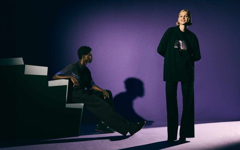 Two fashion models pose in studio with purple background and stairs prop. One model seated, one standing; both wear black attire.