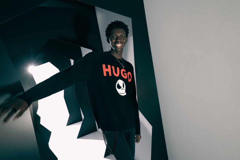Smiling man in a black Hugo sweater with a skull graphic poses in a modern studio with black and white geometric panels and cool lighting.