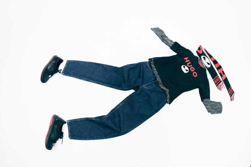 Overhead shot of an apparel flat lay on a white background featuring a dark blue pair of jeans, a black HUGO shirt, and black and red sneakers.