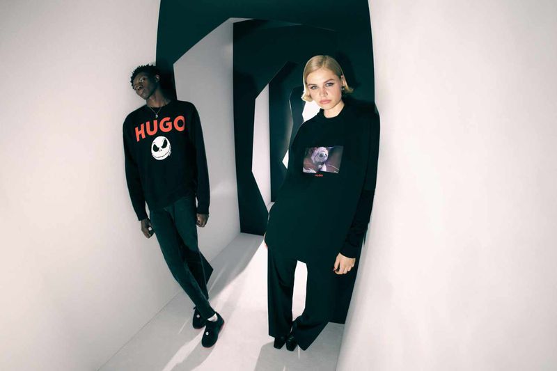 Two models in dark clothing pose in a white corridor, one wearing a Hugo sweatshirt and the other with an image on their shirt.