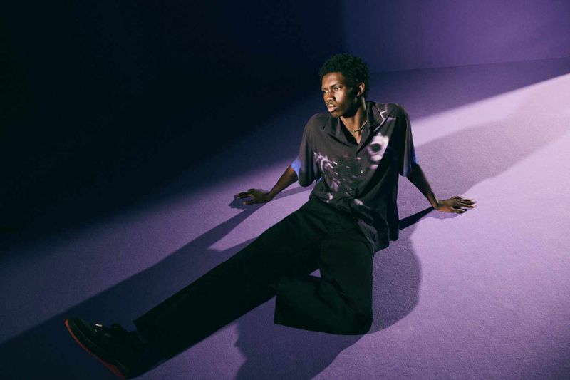 Fashionable African-American adult man in casual graphic shirt and slacks sitting in a room lit with purple light and stark shadows.