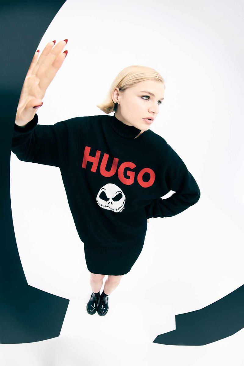Young woman posing in black sweater dress with HUGO logo and cartoon skull on a white backdrop with black geometric shapes.