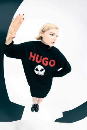 Young woman posing in black sweater dress with HUGO logo and cartoon skull on a white backdrop with black geometric shapes.