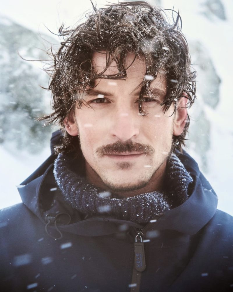 A man with a mustache stands in a snowy environment wearing a navy hooded jacket with snow in his hair for a close-up headshot.