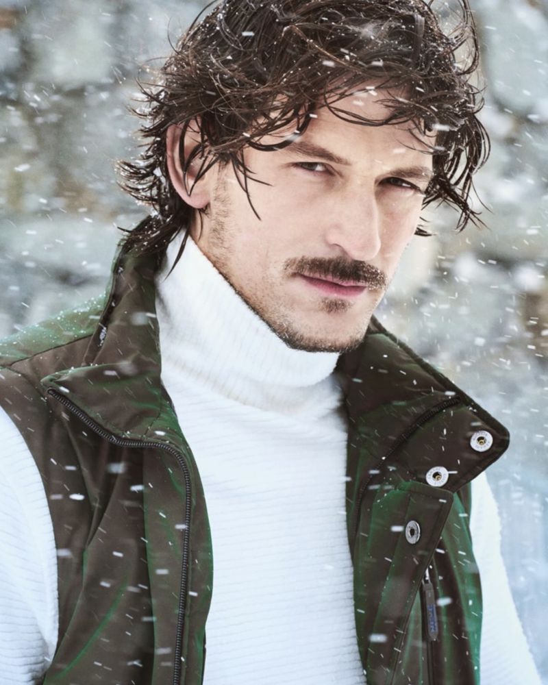 Portrait of a handsome adult male with dark hair and a mustache, wearing a white turtleneck and an olive green vest, in the snow.