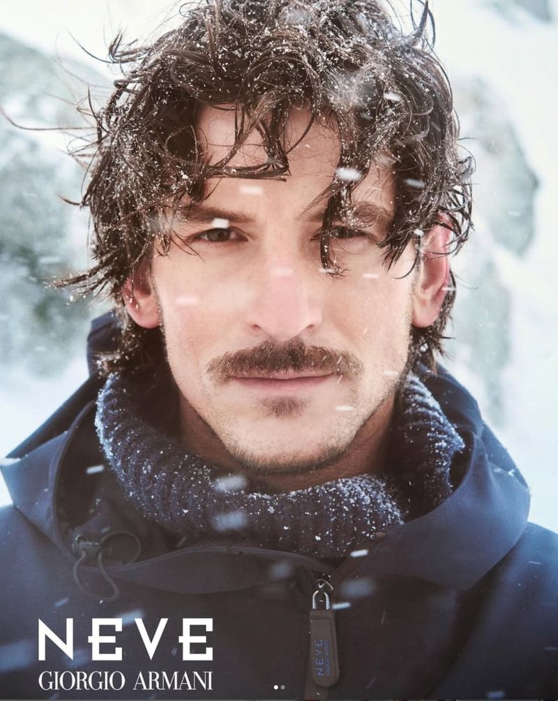 Close-up shot of a man with a moustache, wearing a navy blue jacket and sweater, dusted with snow. 'NEVE GIORGIO ARMANI' is printed.