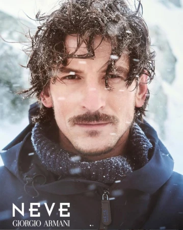 Close-up shot of a man with a moustache, wearing a navy blue jacket and sweater, dusted with snow. 'NEVE GIORGIO ARMANI' is printed.