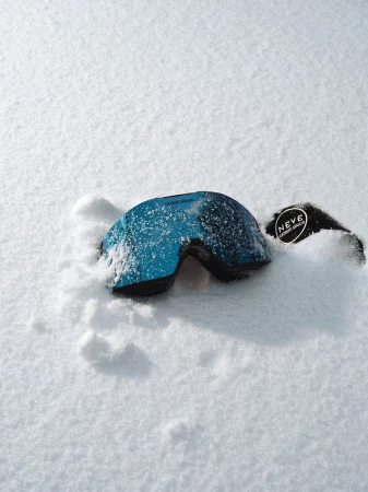 Blue ski goggles covered in snow resting in a snow-covered field with the brand logo visible on the goggle's strap.