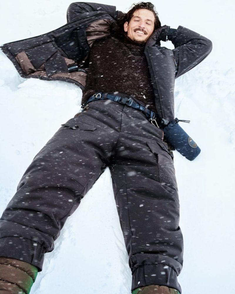 Man lying on his back in the snow, wearing a dark-colored winter jacket, pants, and gloves, with a smile on his face.