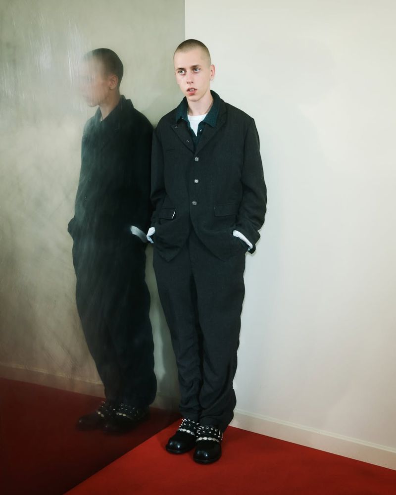 A young adult male in a black suit poses indoors with a serious expression. His reflection is visible in a mirror to his left.