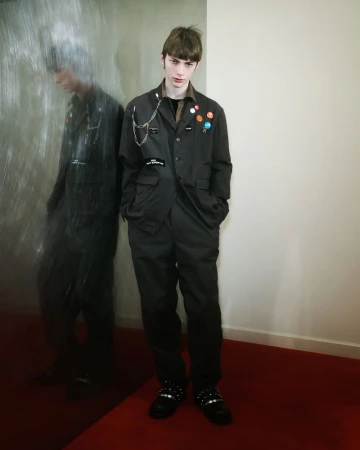 A young adult male stands with a dark outfit decorated with badges and chains, with a muted, neutral background and an elongated reflection.