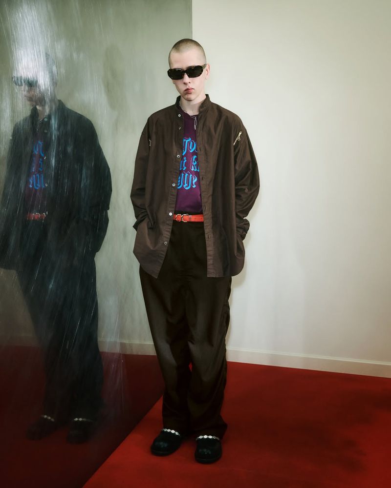 Adult male poses in brown shirt, purple t-shirt, dark pants, and black sunglasses indoors on a red carpet with a partial mirror reflection.