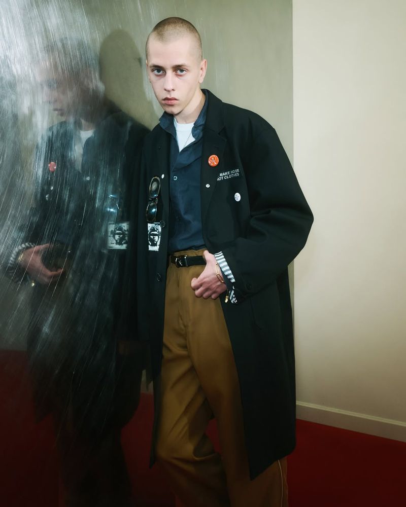 Male fashion model in a black overcoat and tan pants poses in an indoor setting with a mirror creating a reflected duplicate.