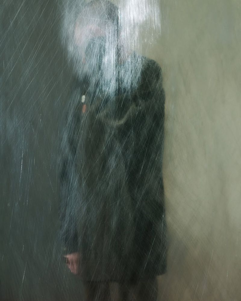 Motion-blurred figure in dark coat stands against mottled wall, rain-streaked effect enhances abstract, artistic style of the photograph.